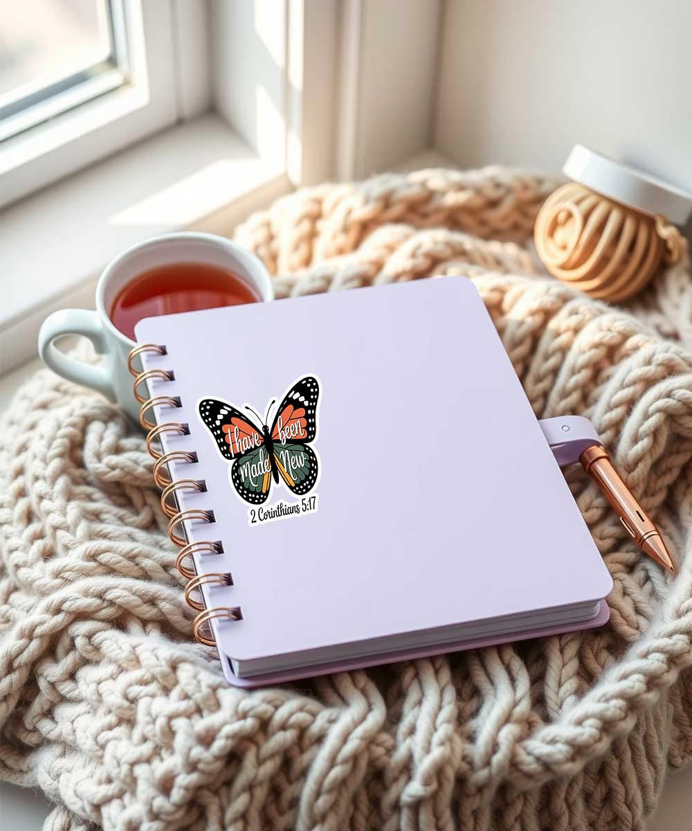 butterfly-christian-sticker-i-have-been-made-new-on-notebook "I have been made New 2 Corinthians 5:17" butterfly design Christian sticker on a light purple notebook, with a knitted blanket and pen.
