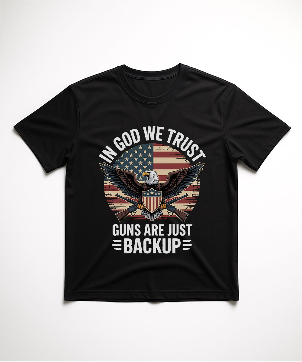 A black t-shirt laid flat, featuring the "In God We Trust, Guns Are Just Backup" design with a patriotic American flag and bald eagle graphic.