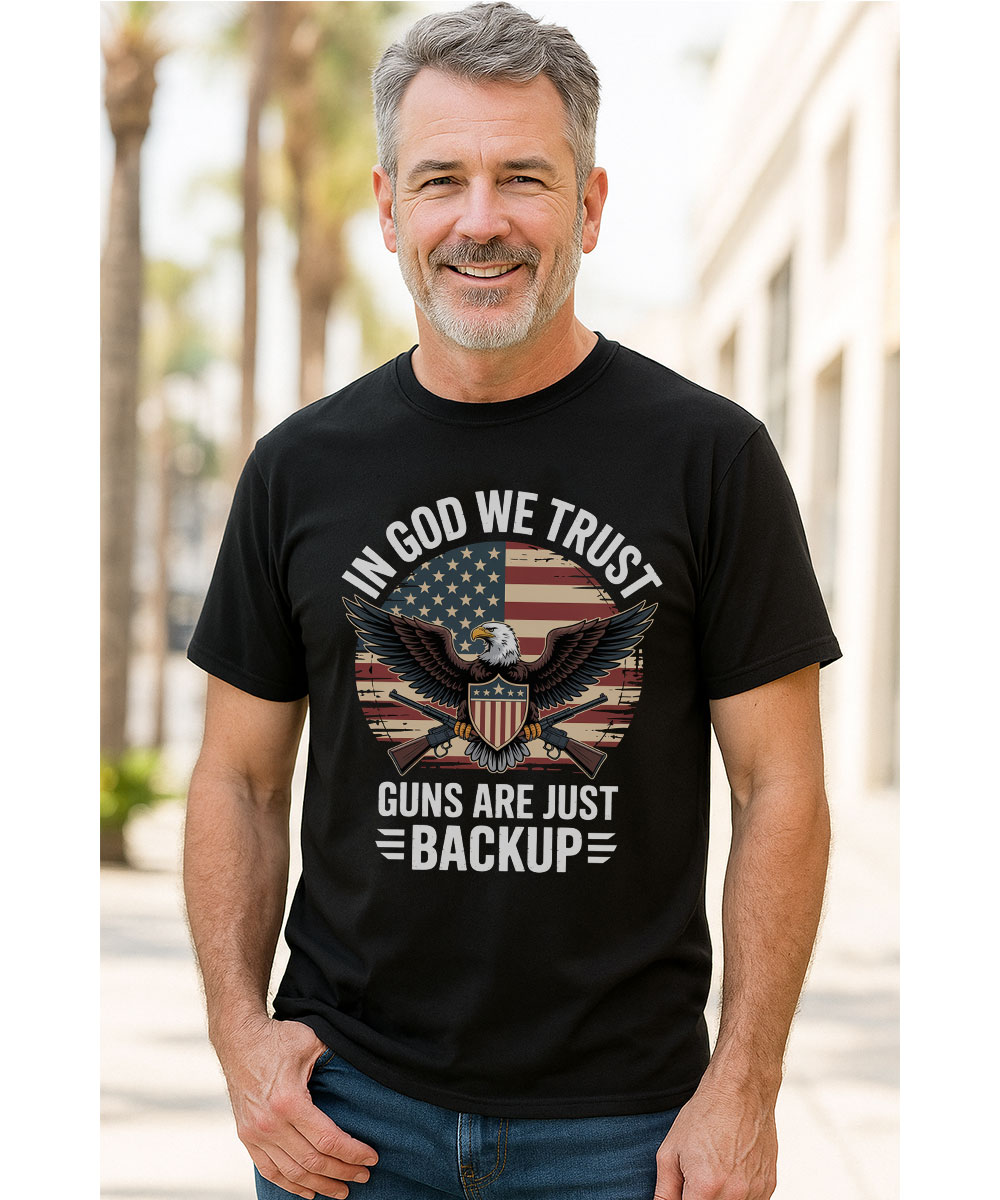 A smiling middle-aged man with graying hair models the "In God We Trust, Guns Are Just Backup" Christian t-shirt outdoors.