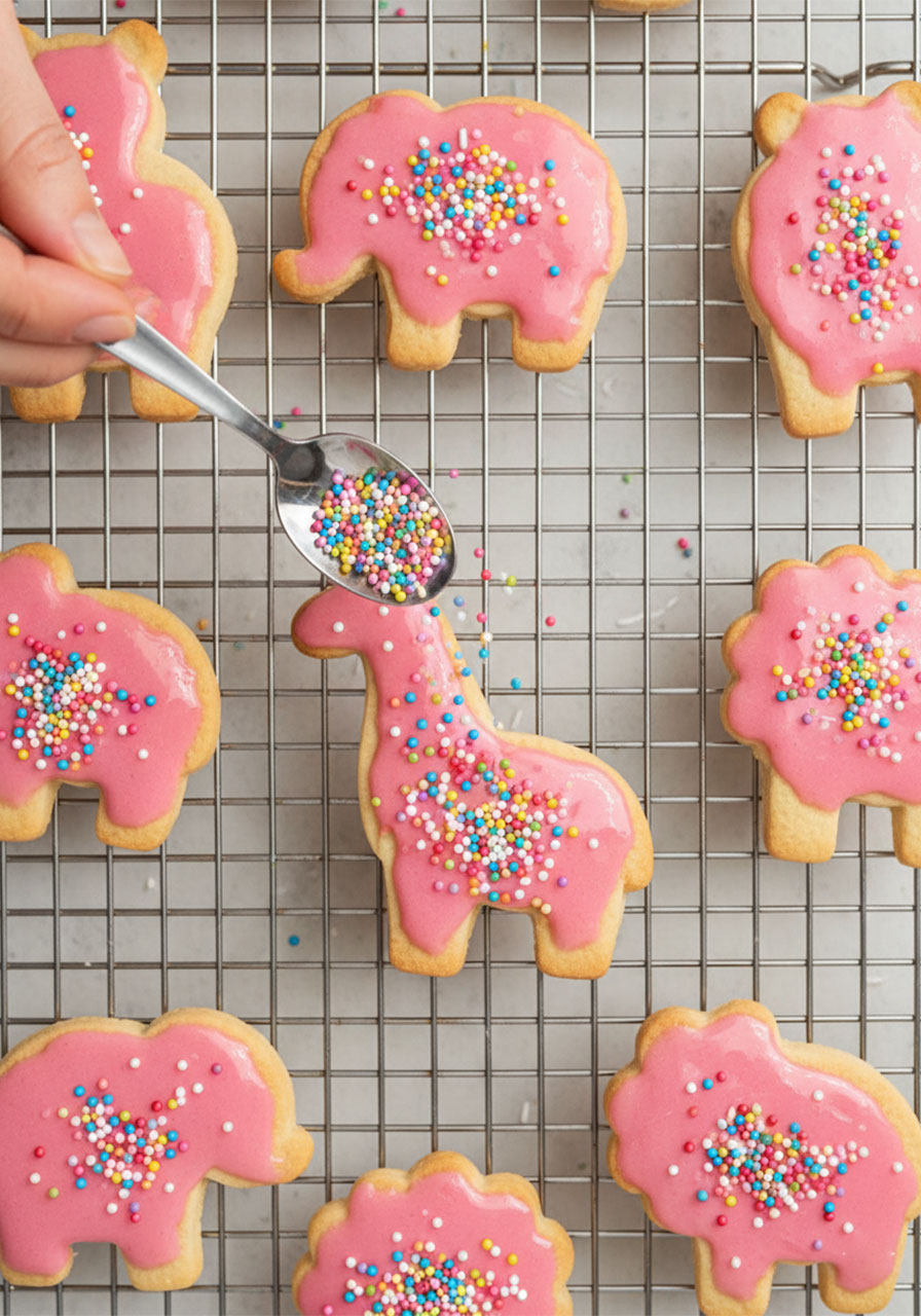 Adding rainbow sprinkles to freshly glazed homemade vegan dairy-free animal cracker cookies on a cooling rack