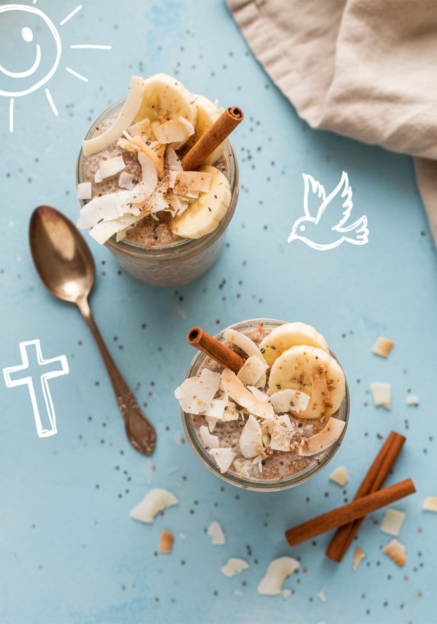 Overhead flat lay of two chai latte chia pudding jars with coconut, banana, and cinnamon sticks on gray surface
