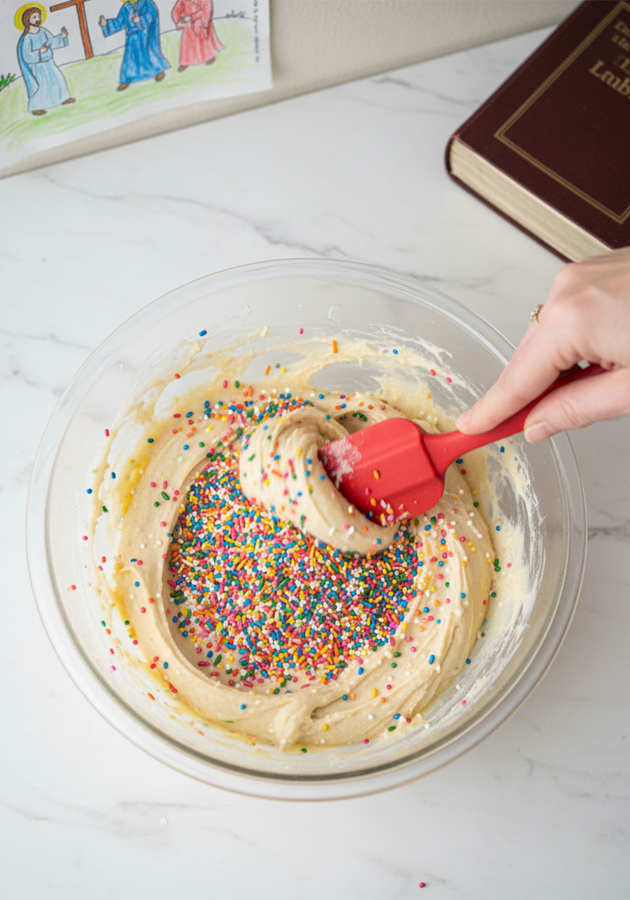 Folding colorful rainbow sprinkles into vegan funfetti sugar cookie dough with a red rubber spatula